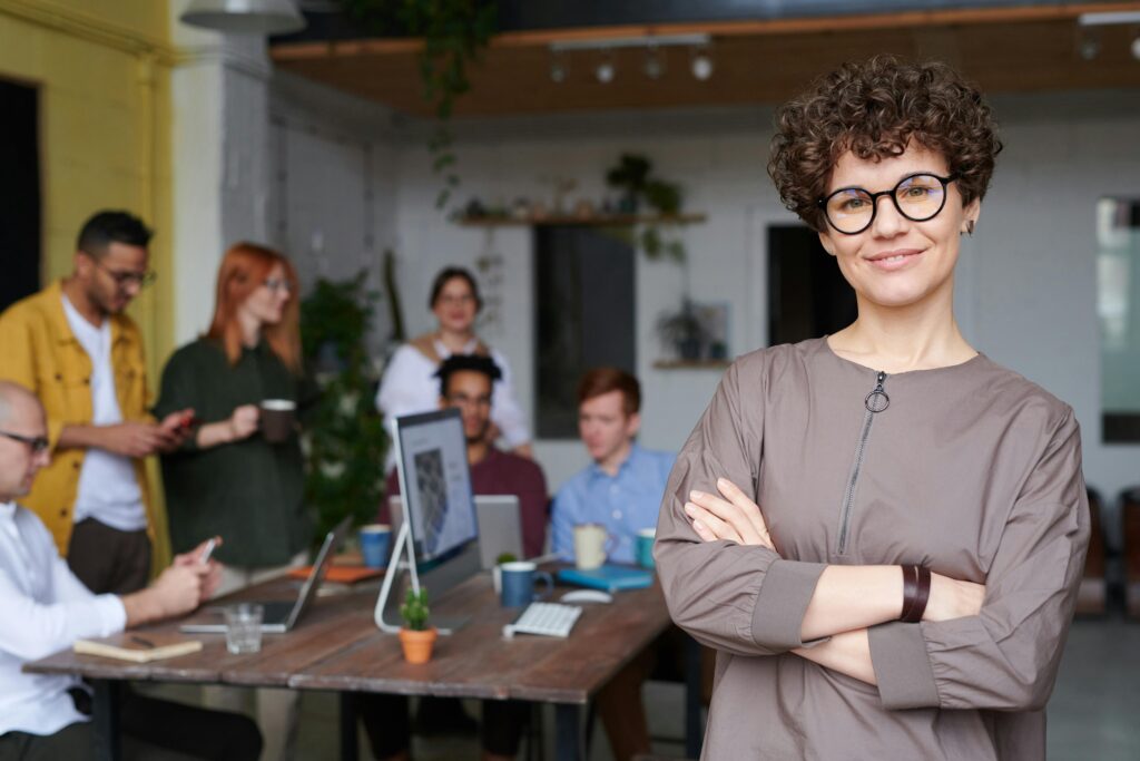 pexels-photo-3184405-3184405-1 Smiling businesswoman with curly hair stands confidently in a modern office space with colleagues.