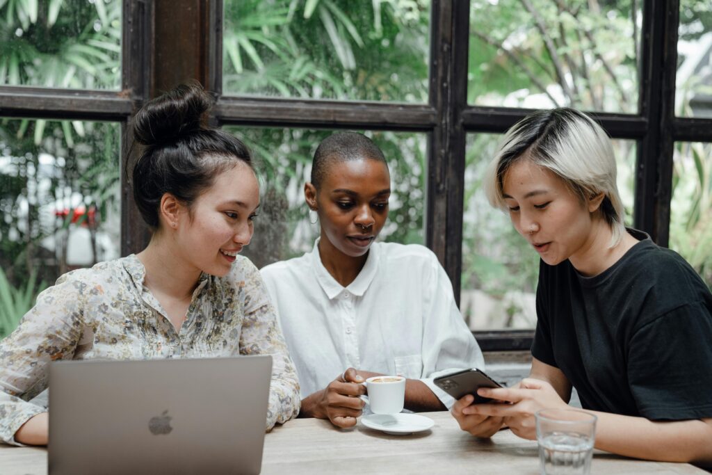 pexels-photo-4350214-4350214 Young Asian and African American women checking posts on social media while sitting in cozy cafe with coffee and laptop near large window with exotic green garden outside